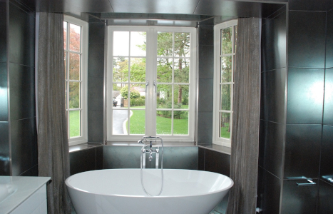 A view of the bath in a black tiled bathroom, with a window overlooking a garden next to the bath.