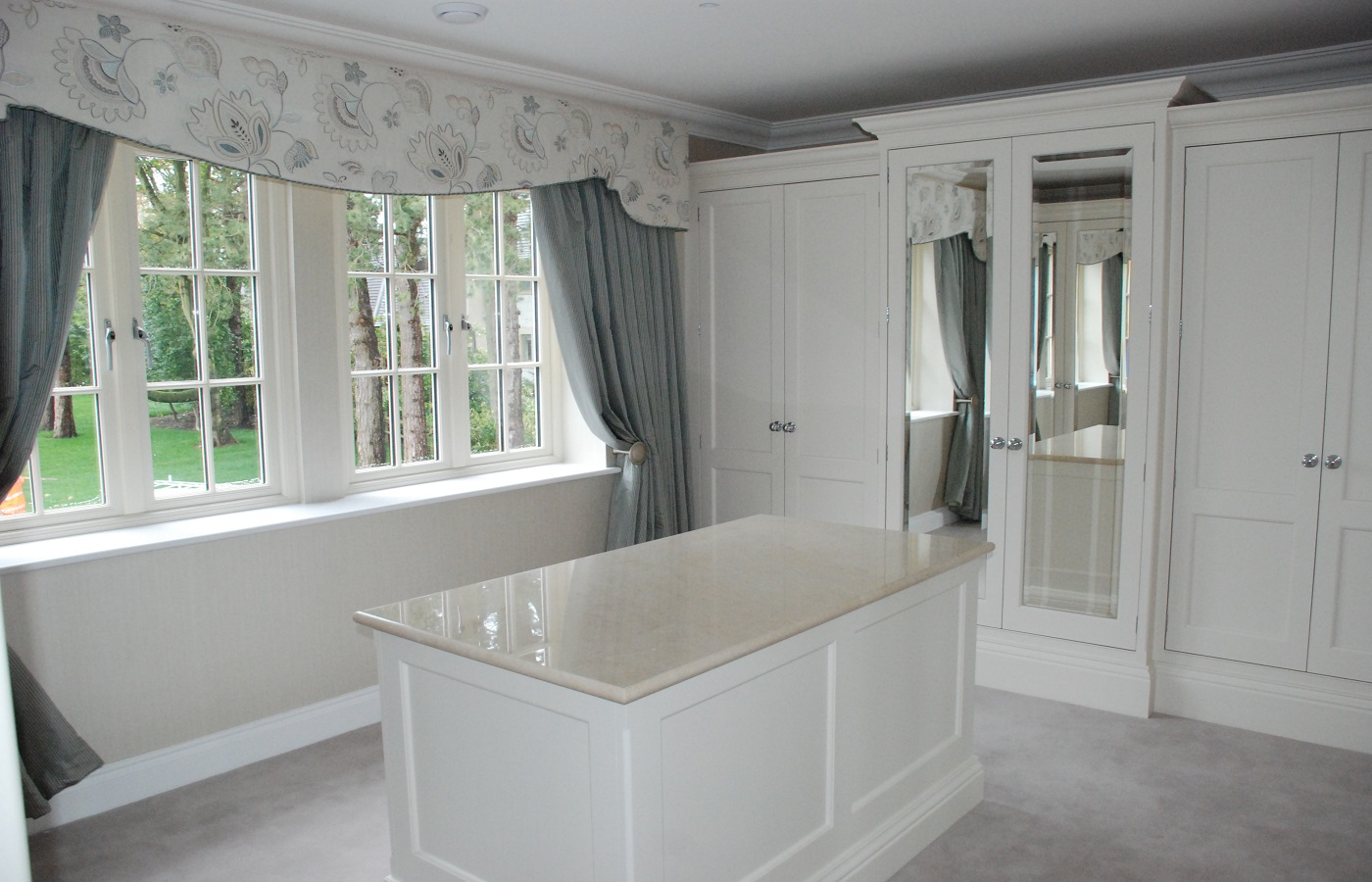 A light white and grey walk in dressing room containing an island and wardrobes, where two doors are mirrored.